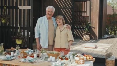 Sıcak, güneşli bir günde, yemek masasının önünde duran, sarılan, gülümseyen, orta yaşlı beyaz kadın ve erkek.