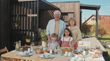 Sıcak güneşli bir günde torunları ve torunlarıyla yemek masasında kameraya bakıp gülümseyen orta yaşlı beyaz kadın ve erkek.