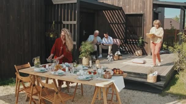 在阳光明媚的夏日 两名年轻的老年白人妇女在户外的木制避暑别墅上桌 她们把装有食物的碗放在桌子上 两名男子坐在扶手椅上 — 图库视频影像