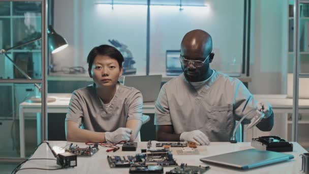 アジア系の女性とアフリカ系アメリカ人の男性の肖像画 手袋や制服を着た男が研究室の机の上に座り カメラのためにポーズをとりながら技術エンジニアとして働く — ストック動画