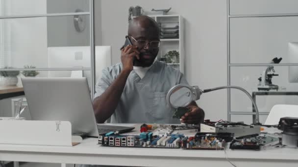 African American Male Engineer Speaking Mobile Phone Examining Chip Magnifying — Stock Video