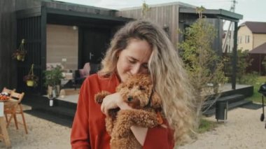 Uzun saçlı, turuncu tişörtlü, sevimli Maltipoo köpeğini tutan, dışarıda kameraya bakıp gülümseyen beyaz bir kadın.