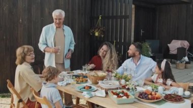 Orta uzunlukta beyaz saçlı beyaz bir adam güneşli bir yaz haftasonu evinin önünde oturup gülümseyen ve konuşan aile üyeleriyle tost yapıyor.