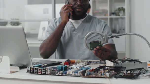 携帯電話で話しているアフリカ系アメリカ人の電子工学者のショットを傾け 研究室の机で働いている間に虫眼鏡でチップを調べる — ストック動画