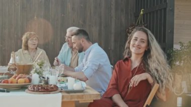 Uzun saçlı, beyaz kadın gülümseyen ve kameraya bakan güneşli yaz gününde dışarıda yemek masasında oturan aile üyeleri arka planda sohbet eden.