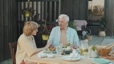 Yazlık evin arka bahçesindeki masada oturan, güneşli bir günde konuşan ve gülümseyen son sınıf beyaz heteroseksüel çift.