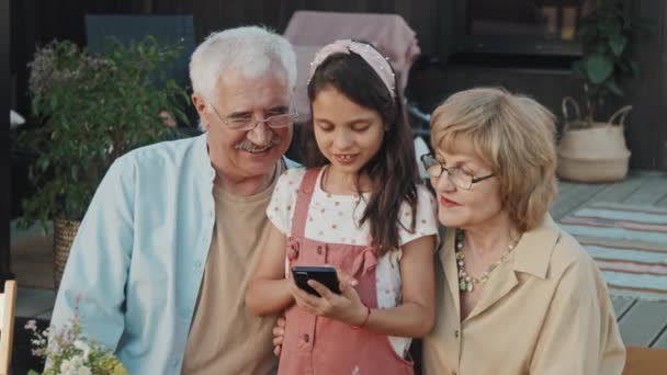 幸せなシニア白人女性と男性の中で彼らのかわいい孫娘を見ますスマートフォンを使用して 晴れた日に夏の家の外に座って 話して笑顔 — ストック動画