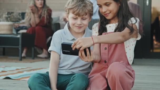 在温暖的夏日 白人小男孩和他的姐姐坐在楼梯间的门廊上 用智能手机自拍 做鬼脸 父母在背景上聊天 — 图库视频影像