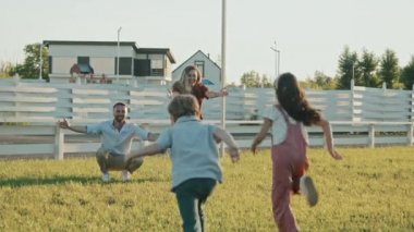 Küçük kız ve oğlanın küçük, mutlu beyaz bir kadın ve erkeğin kollarına koşması, sıcak bir yaz günü arka bahçede yeşil çimlerin üzerinde kucaklaşmaları.
