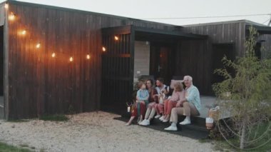 Yazlık evin verandasında oturan, konuşan ve gülümseyen beyaz bir çift, iki çocuk ve köpek.