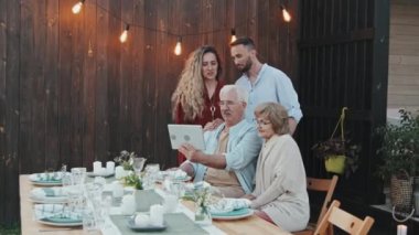 Orta uzunlukta mutlu, genç, beyaz bir çift yaz akşamı arka bahçede masada oturan yaşlı kadın ve adamın arkasında duruyor. Tablet bilgisayardan arıyorlar.