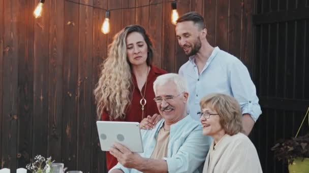 若い白人女性と男性の中で夏の夜に裏庭に座っている老夫婦の後ろに立って ビデオはタブレットコンピュータを介して呼び出し — ストック動画
