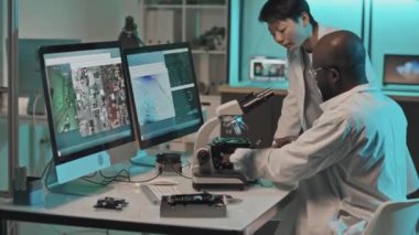 Beyaz önlüklü çok ırklı teknik mühendislerin mikroskopla bağlantılı görüntüleri tartışırken laboratuvarda elektronik bileşenleri incelerken görüntülerini yukarı kaldır.