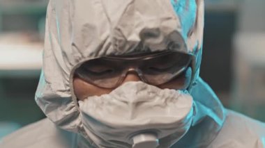 Örtülü, maskeli ve gözlüklü Asyalı kadın laboratuvar çalışanı portresi başını kaldırıp kameraya bakıyor.