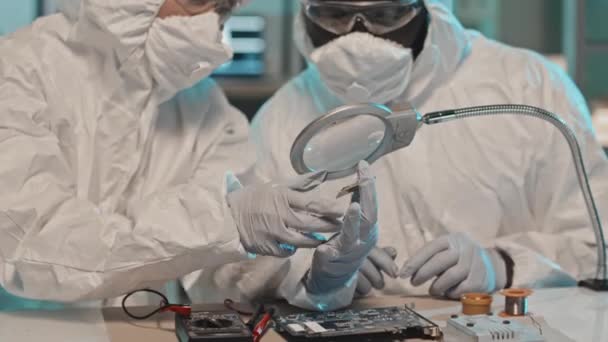虫眼鏡の下でコンピュータチップを見て 研究室で協力しながら議論をしながら 全体的なスーツ マスクや手袋で2人の多様なエンジニアのショットを傾ける — ストック動画