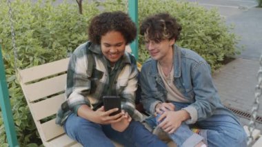 Orta uzunlukta bir zenci kız akıllı telefon kullanıyor, gözlüklü ve kot ceketli neşeli beyaz adamla konuşuyor, yaz sabahının erken saatlerinde salıncakta oturuyor, gülümsüyor.