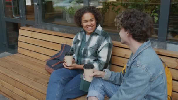 夏日早晨 中等身材的黑人女孩坐在咖啡店外的长椅上 白人男性朋友穿着牛仔夹克 喝着纸杯咖啡 谈着话 面带微笑 — 图库视频影像