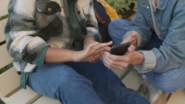 Kot giyen, bacaklarıyla açık havada sallanan ve yazın akıllı telefon kullanan iki üniversite öğrencisinin yüksek açılı görüntüsü.