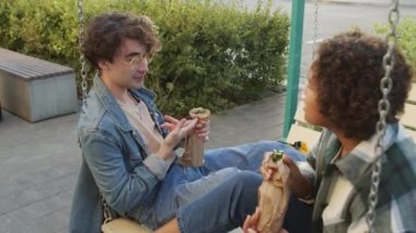 Orta uzunlukta, kot ceket giyen, Afrikalı Amerikalı bir kadınla konuşan, yazın salıncakta sallanan, kağıt torbalardaki şişelerden bira içen mutlu bir beyaz adam.
