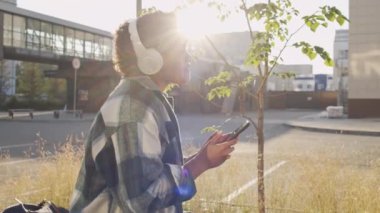 Afrika kökenli Amerikalı kadın Gen Zer 'in arka planda oturduğu, kulaklık taktığı, akıllı telefon kullandığı, güneşli yaz günlerinde müzik dinlediği