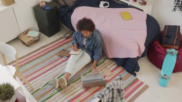 非洲裔美国女大学生穿着休闲装 白天坐在卧室的地板上 用笔记本电脑上网 用笔记本记笔记 — 图库视频影像