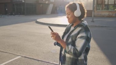 Afrika kökenli Amerikalı bir kızın şehir dışında dikilirken, kulaklık takarken, akıllı telefon kullanırken, güneşli yaz günlerinde müzik dinlerken arka plandaki görüntüsü.