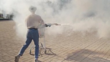 Neşeli beyaz erkek üniversite öğrencisi, güneşli yaz gününde şehirde sis bombası tutan dikkatsiz siyahi bir kızla alışveriş arabasını itiyor.