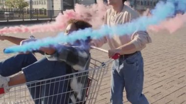 Neşeli beyaz erkek üniversite öğrencisi, güneşli yaz gününde şehirde elinde kırmızı ve mavi duman bombası tutan dikkatsiz siyahi bir kızla alışveriş arabasını itiyor.