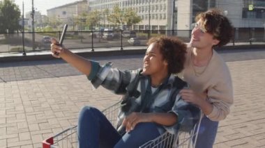 Orta uzunlukta mutlu bir siyah kız şehirde alışveriş arabasında oturuyor, neşeli beyaz erkek arkadaşıyla selfie çekiyor, gülümsüyor, eğleniyor, güneşli bir günde surat yapıyor.