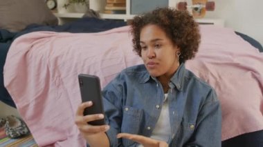 Yirmi yaşındaki siyahi bir kızın, parlak ve temiz yatak odasında sırtüstü uzanmış, gündüz vakti video görüşmesi için akıllı telefonu kullanmasının orta boy yakın çekimi.