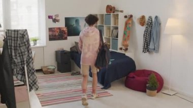 Kıvırcık, genç, beyaz bir adam yatak odasına geri dönüyor, uzun tahta ve sırt çantası bırakıyor, yatağa düşüyor ve gündüzleri dinleniyor.