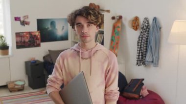 Kıvırcık saçlı beyaz erkek üniversite öğrencisinin portresi. Kapüşonlu, taşınabilir bilgisayarlı, gündüz yatak odasında kameraya bakıyor.