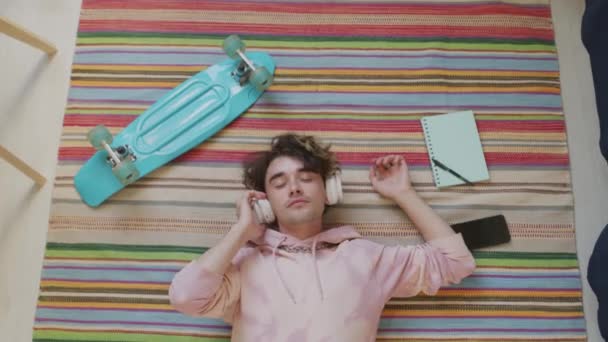 昼間に家の床にカーペットの上に横たわって スマートフォン プラスチック製のスケートボード ペンとノートブックに囲まれて パーカーと耳の上のヘッドフォンを身に着けている白人の若い男のトップビュー — ストック動画