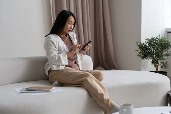 Молодая азиатская бизнесвумен сидит на диване, прокручивая в мобильном телефоне — стоковое фото