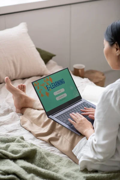 Günümüz genç bayan gündelik kıyafetleriyle online ders sırasında yatakta oturuyor.