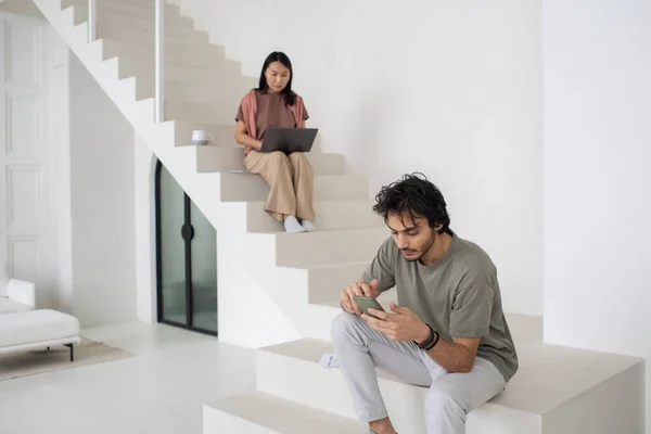 Akıllı telefonlu genç beyaz adam dizüstü bilgisayarlı Asyalı bir kadına karşı oturuyor. — Stok fotoğraf