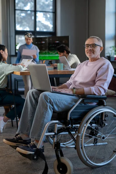 Rijpe werknemer met laptop op zijn knieën zittend in rolstoel — Stockfoto