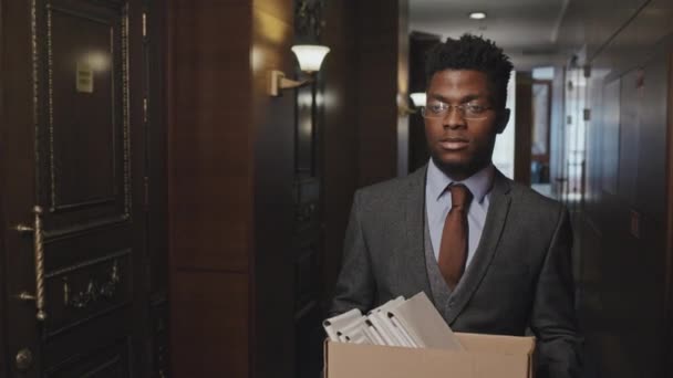 アフリカ系アメリカ人のビジネスマンが解雇された後オフィスを出る際に廊下を通ってファイルフォルダで満たされた箱で歩く — ストック動画