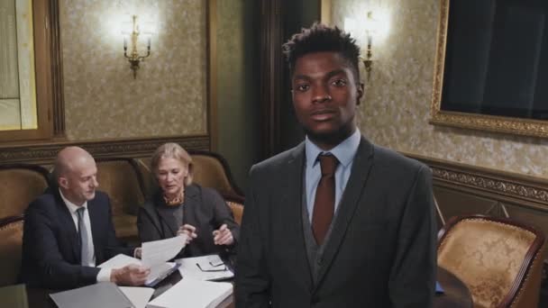 若いアフリカ系アメリカ人弁護士の肖像がオフィスに立ってカメラにポーズをとっているのに対し 白人の同僚はバックグラウンドで働いている — ストック動画