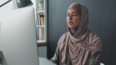 Başörtülü genç Müslüman kadın ofis masasında oturmuş bilgisayardaki video görüşmesinde belgeleri tartışıyor.