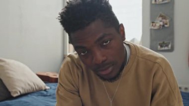 Genç Afrikalı Amerikalı bir adamın portresi başını kaldırıyor ve yatak odasında otururken kameraya poz veriyor.