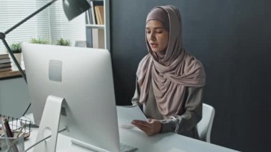 Başörtülü güzel Müslüman bir kadının internet üzerinden ders veya iş görüşmesi yaparken ofis masasında oturup bilgisayar üzerinden kağıt tartışması.