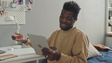 Kablosuz kulaklıklı genç Afrikalı Amerikalı adam evde oturuyor, gülümsüyor ve dijital tablet üzerinden çevrimiçi video konuşmaları yapıyor.