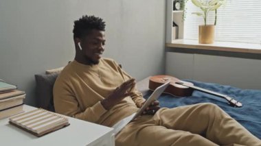 Genç Afrikalı Amerikalı adam evde yatağında dinlenirken dijital tablette gülümseyip sohbet ediyor.