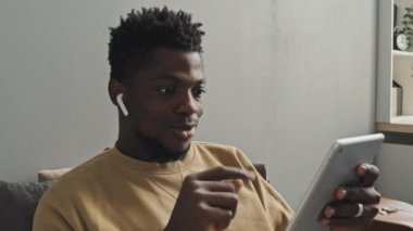 Kablosuz kulaklıklı, gülümseyen, el sallayan ve dijital tablette konuşan Afro-Amerikan bir adam.