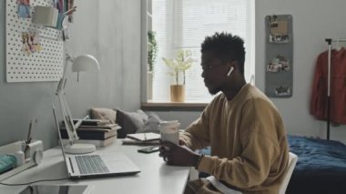 Kablosuz kulaklıklı Afrikalı Amerikalı bir adam kahve fincanı ile masada oturuyor ve evdeki dizüstü bilgisayarında online video görüşmesi yapıyor.