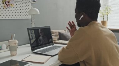 Kablosuz kulaklıklı Afro-Amerikalı bir adam evde online ders alırken, masa başında oturmuş video aracılığıyla tesettürlü bir kadınla konuşuyor.