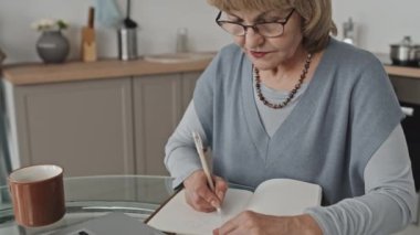 Kıdemli bir kadının not yazıp evde uzaktan öğrenirken online ders izlediği bir kare.