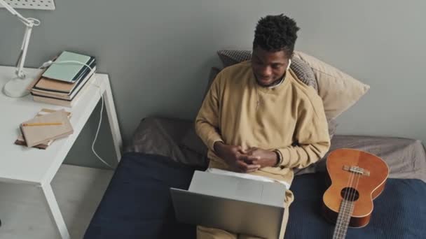 高角度ショットのアフリカ系アメリカ人男性で無線イヤフォンに座っているギターでベッドの上に何かを議論し ラップトップ上のオンラインビデオ通話 — ストック動画