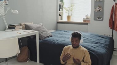 Afrika kökenli Amerikalı bir adamın kablosuz kulaklıklarıyla yatak odasında otururken ve evde e-öğrenim görürken bilgisayardaki video konuşmalarını aşağı eğ.